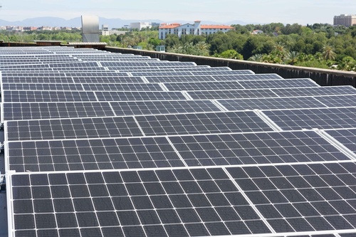 Un grand ensemble de panneaux solaires s'étend sur un toit, avec un paysage urbain et verdoyant visible à l'arrière-plan sous un ciel clair.