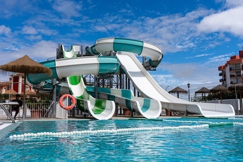 Toboganes de agua verdes y blancos se elevan sobre una piscina azul de un parque acuático, con edificios y una sombrilla de paja en el fondo bajo un cielo azul con nubes.