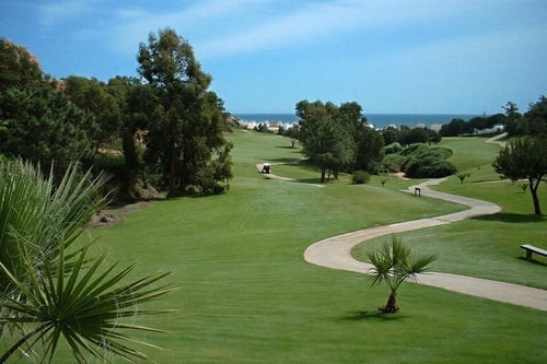 A picturesque golf course featuring lush green fairways, scattered trees, and a winding cart path leads towards the distant ocean under a clear blue sky.