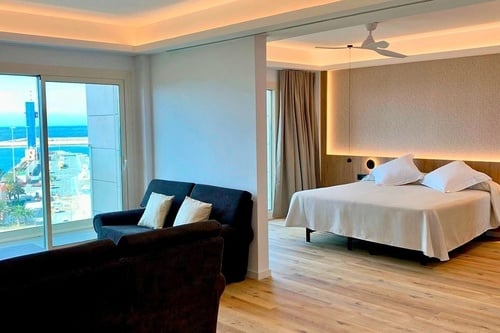 Une chambre d'hôtel moderne et lumineuse offre un espace salon avec des canapés et une chambre séparée, le tout avec une vue imprenable sur la mer et un port.