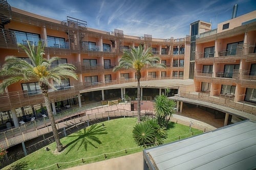 La imagen muestra un complejo hotelero curvado de color melocotón con múltiples balcones y barandales de madera, que rodea un patio interior con césped verde y palmeras bajo un cielo azul con nubes dispersas.