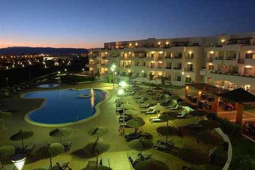 An illuminated resort complex with a large swimming pool, numerous lounge chairs under straw umbrellas, and a multi-story building with lighted balconies is captured at dusk.