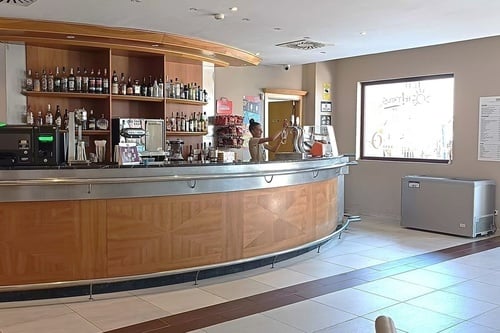 En un bar, una camarera sirve una cerveza detrás de una barra bien surtida con botellas y máquinas de café.