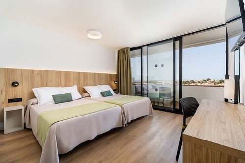 Una habitación de hotel moderna y luminosa con dos camas individuales, suelo de madera y un amplio ventanal que da a un balcón con vistas.