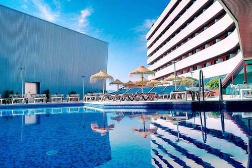 Una piscina exterior de baldosas azules refleja un edificio hotelero moderno y otro industrial de color azul bajo un cielo despejado, flanqueada por tumbonas y sombrillas de paja.