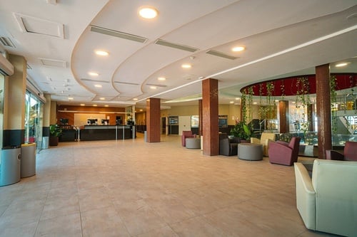 A bright and spacious modern hotel lobby features a long reception desk, multiple comfortable seating areas with armchairs and ottomans, decorative tiled pillars, and a sleek ceiling with recessed lighting.