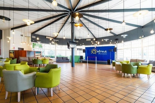 Un amplio y luminoso salón de hotel o resort con un techo de diseño único, múltiples lámparas colgantes, asientos de diferentes colores, un suelo de baldosas y un escenario con telón azul.