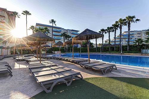 Vista da piscina do hotel com espreguiçadeiras e guarda-sóis de palha, com o sol a brilhar através das palmeiras. Hotel tudo incluído em La Pineda | Ohtels La Hacienda