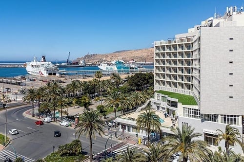 Une vue aérienne présente un port méditerranéen animé avec des ferries, un hôtel moderne entouré de palmiers, et une route fréquentée, sous un ciel bleu clair.