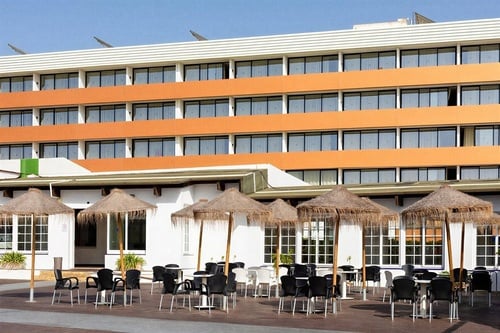Una imagen muestra un hotel moderno con una fachada naranja y blanca, que cuenta con una terraza exterior amueblada con mesas, sillas y sombrillas de paja.