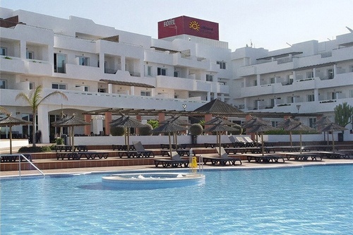 A large white multi-story hotel building with balconies overlooks a swimming pool with a built-in hot tub, surrounded by sun loungers and straw umbrellas.