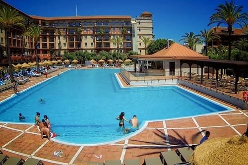 A large, sunny outdoor swimming pool at a resort is bustling with people enjoying the water and lounging by the poolside, with a multi-story hotel building and palm trees in the background.
