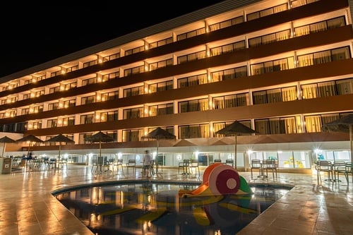 Un hotel con todas sus habitaciones iluminadas por la noche, con una piscina infantil y un área de estar exterior en primer plano.