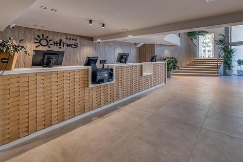 Une réception d'hôtel moderne avec un long comptoir en bois texturé, des écrans d'ordinateur et un grand escalier menant à l'étage est visible dans cette image.