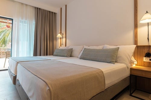 Dormitorio de hotel de estilo moderno con dos camas juntas, cortinas neutras y un balcón soleado con palmeras al fondo.