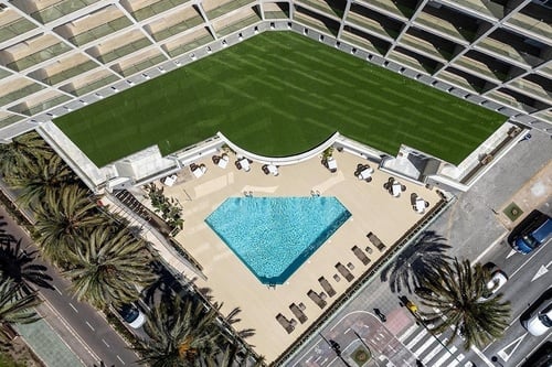 Une vue aérienne montre un complexe moderne avec un toit-terrasse en gazon, une piscine en forme de diamant entourée de chaises longues et de tables, ainsi que des palmiers bordant une rue adjacente.