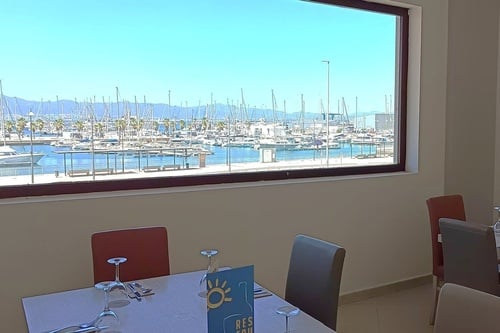 Una vista desde un comedor de un restaurante con una gran ventana que da a un puerto deportivo lleno de veleros y barcos, con montañas de fondo bajo un cielo azul.