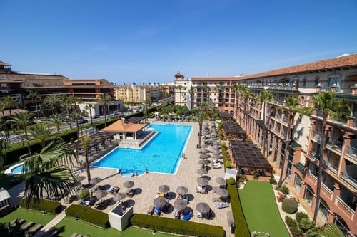 This is an elevated view of a vibrant hotel resort featuring a large, blue swimming pool surrounded by sun loungers and palm trees, with adjacent buildings under a clear sky.