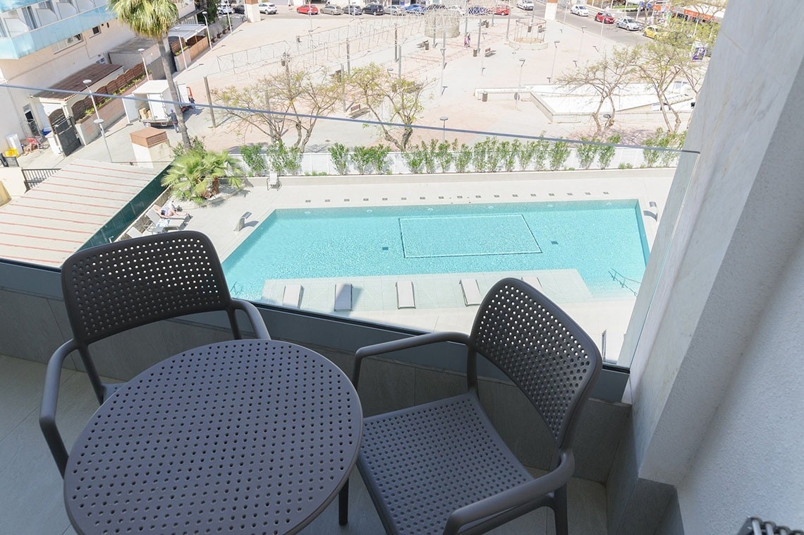 Desde un balcón amueblado, se observa una piscina y un entorno urbano con calles y edificios al fondo.