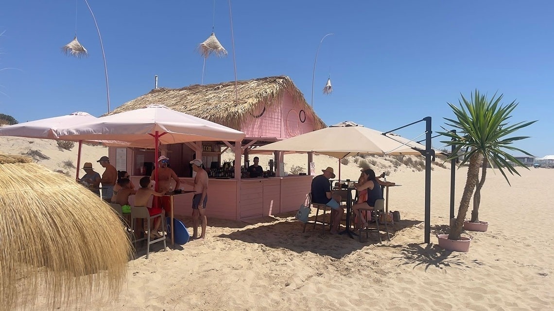un grupo de personas están sentadas frente a una cabaña de paja rosa en la playa