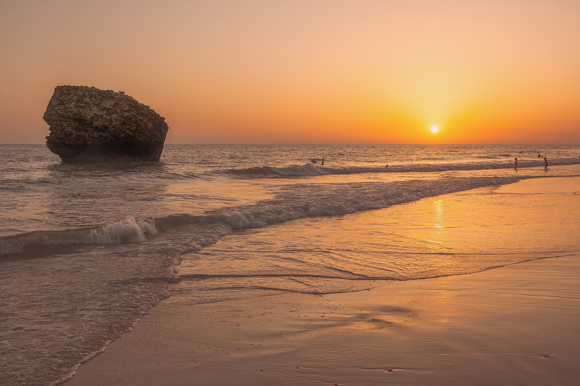 Atardecer vibrante en playa con sol naranja sobre el mar, gran roca y bañistas. Destino de vacaciones costero.