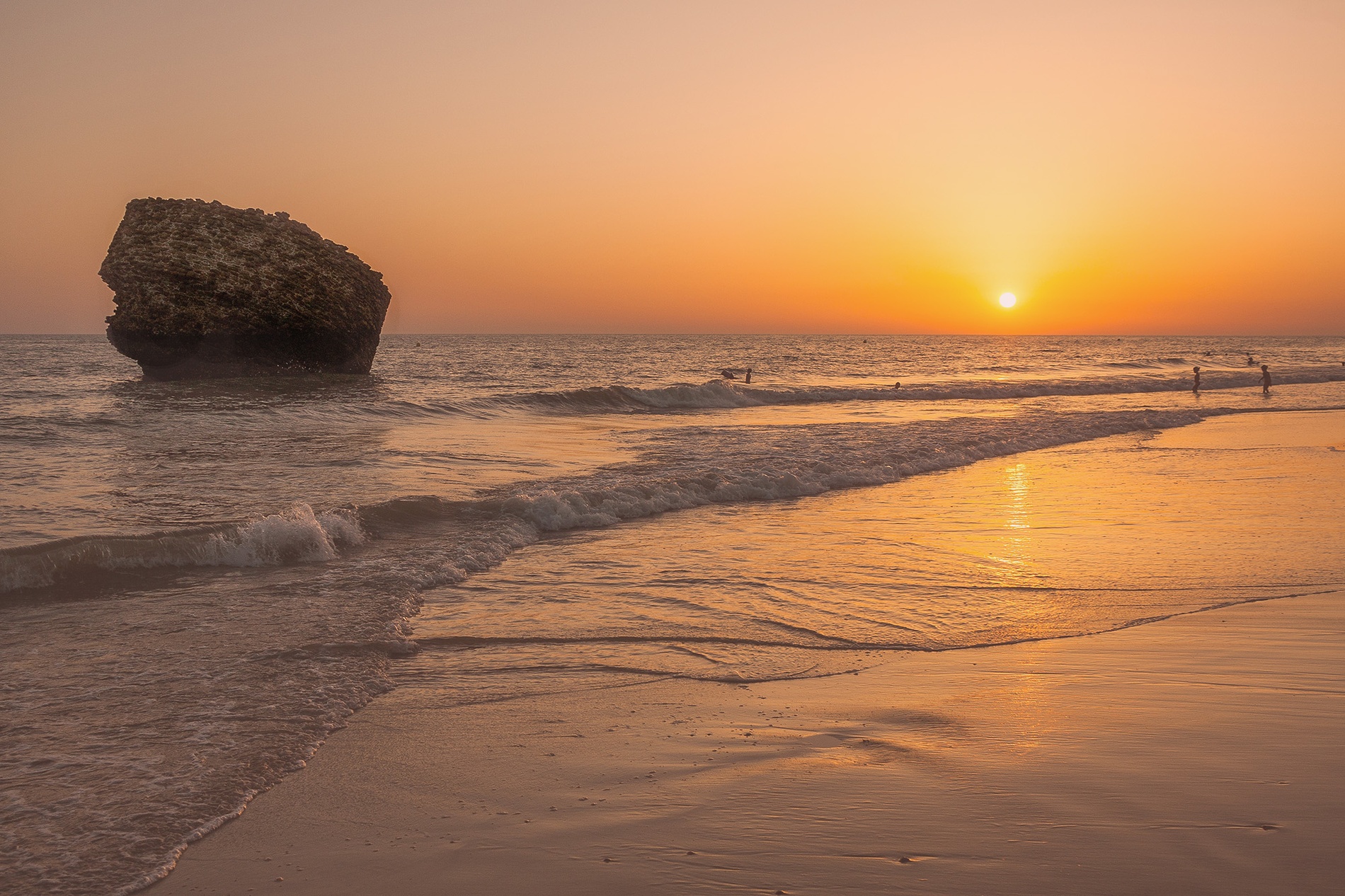 Atardecer vibrante en playa con sol naranja sobre el mar, gran roca y bañistas. Destino de vacaciones costero.