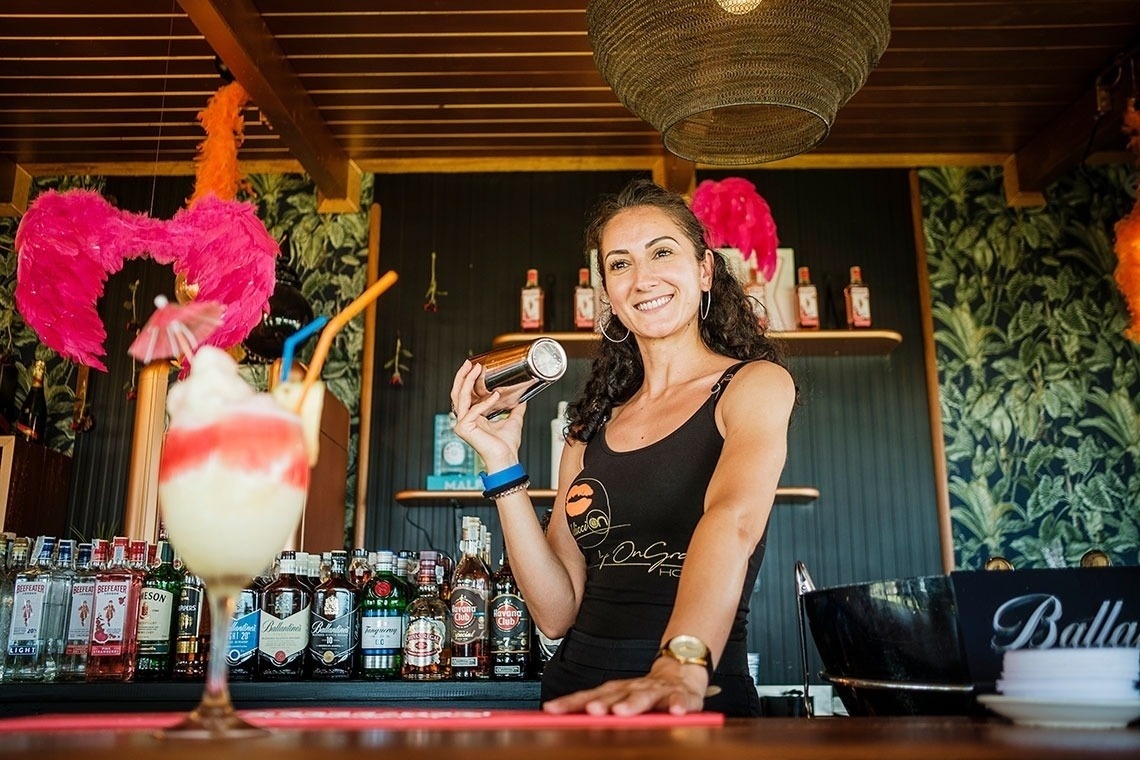 a woman standing behind a bar holding a shaker next to a ballantine bottle