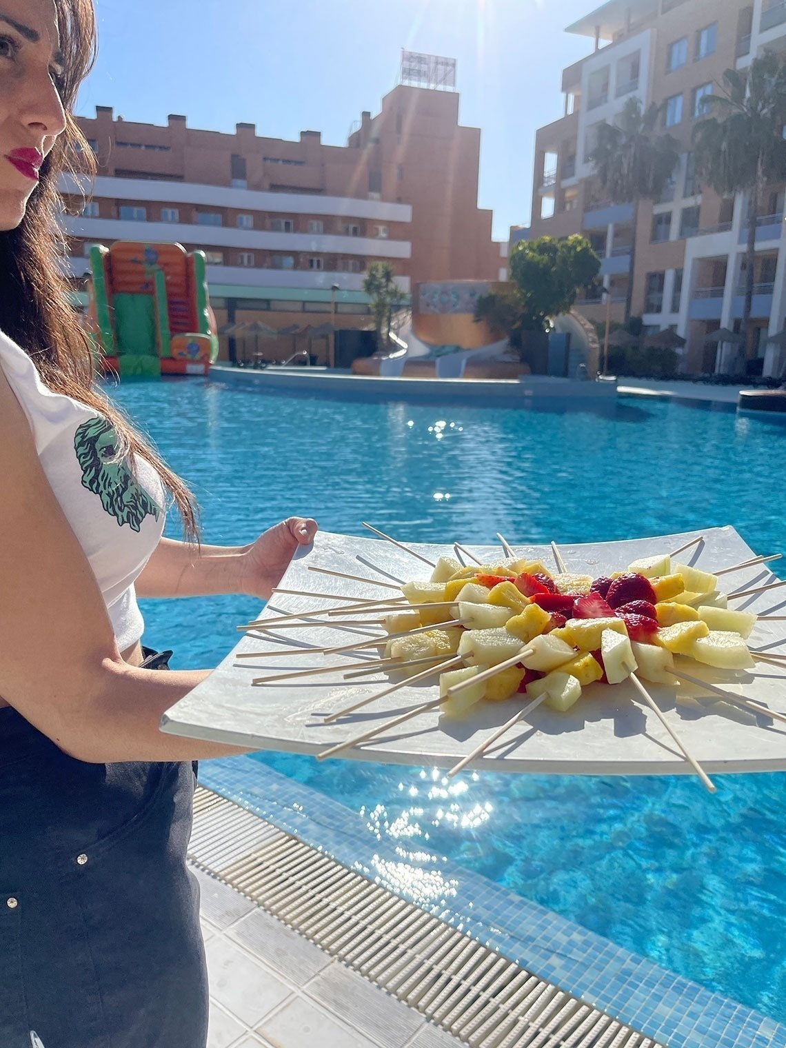 una mujer sostiene un plato de frutas en palillos frente a una piscina