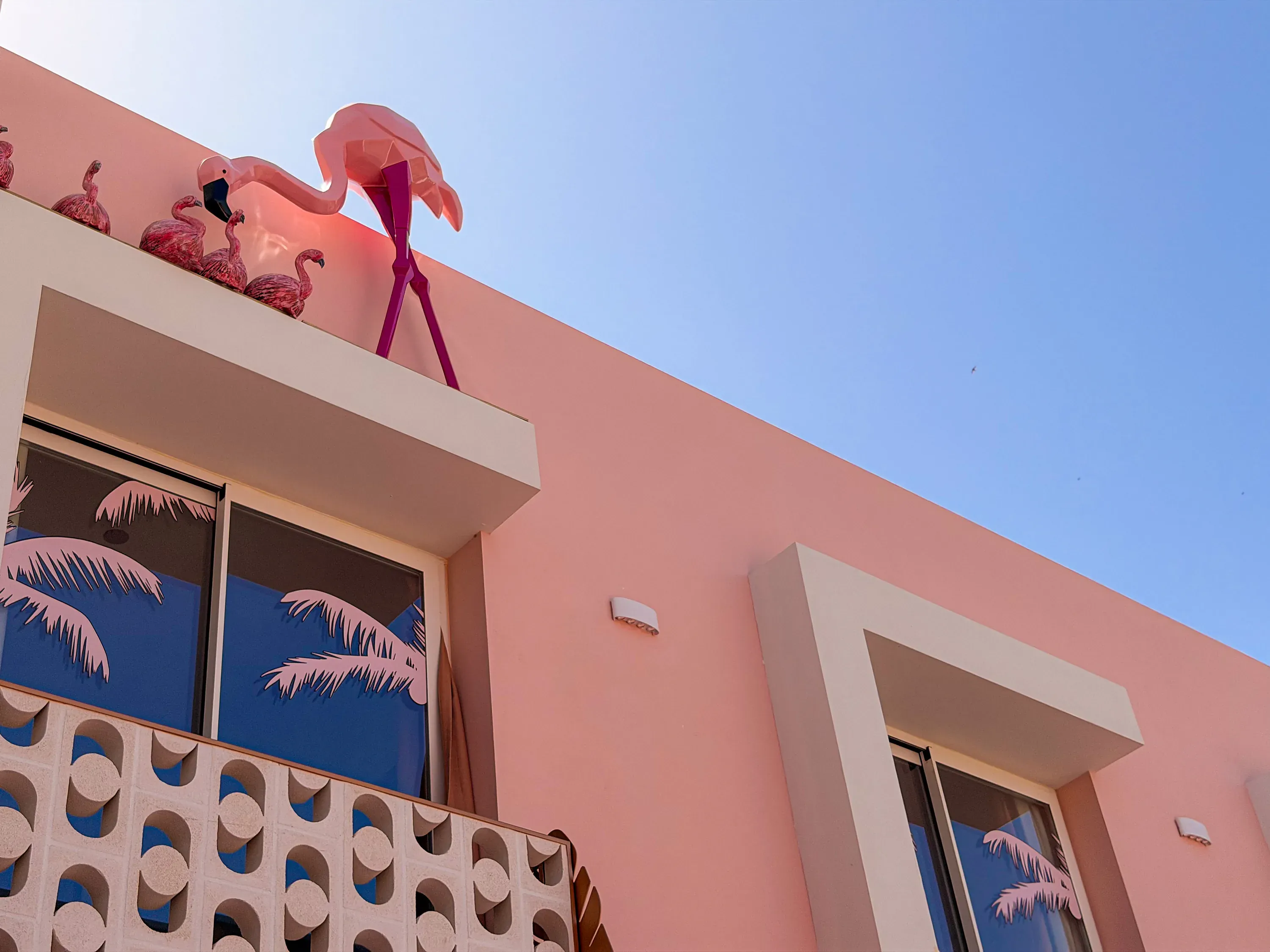 Fachada rosa de hotel con flamencos decorativos y palmeras en las ventanas, ideal para tu estancia ON Pink Motel | Matalascañas.