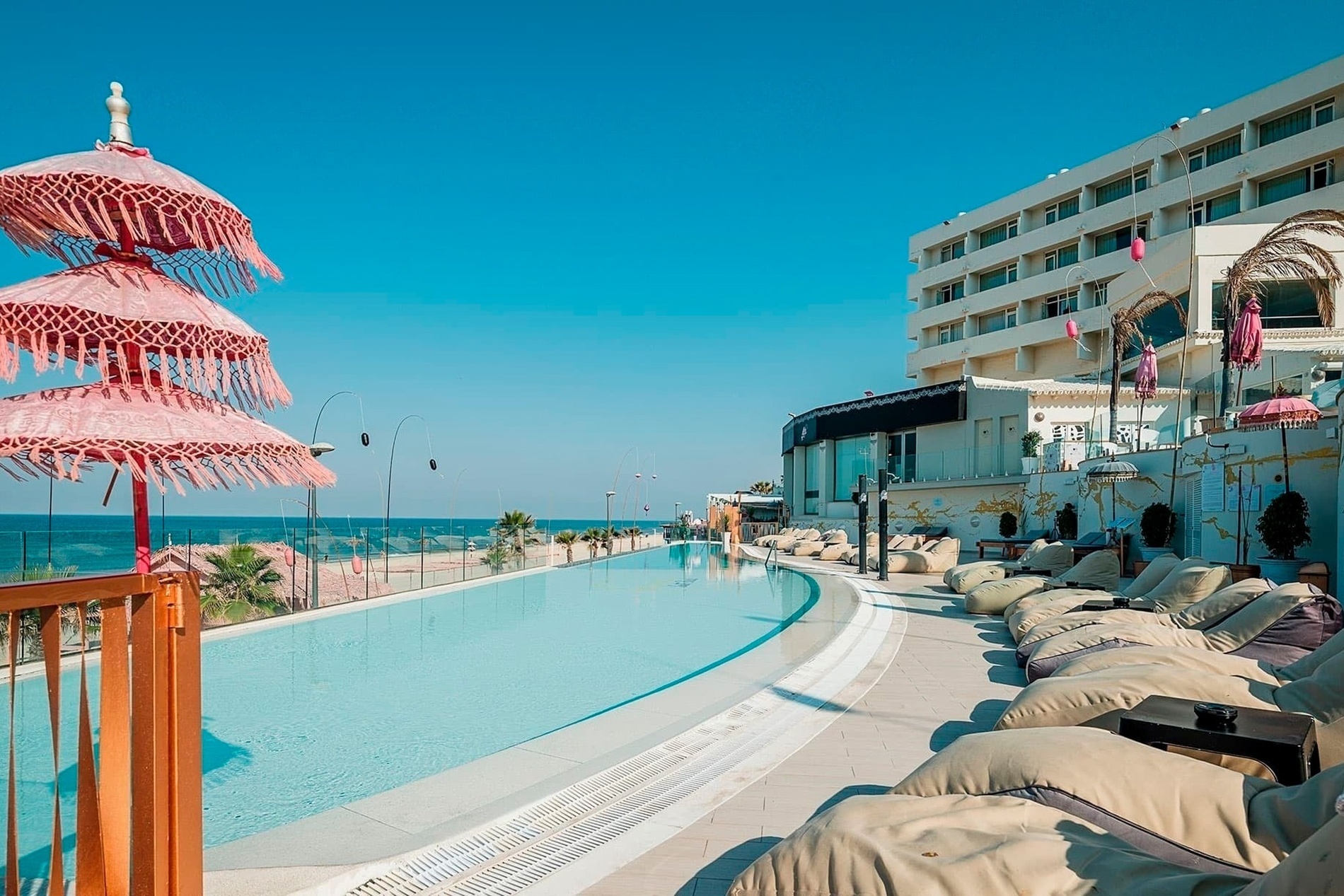 Una lujosa piscina infinita se extiende frente al mar, adornada con sombrillas rosadas y cómodas tumbonas, con un hotel moderno de fondo bajo un cielo azul claro.