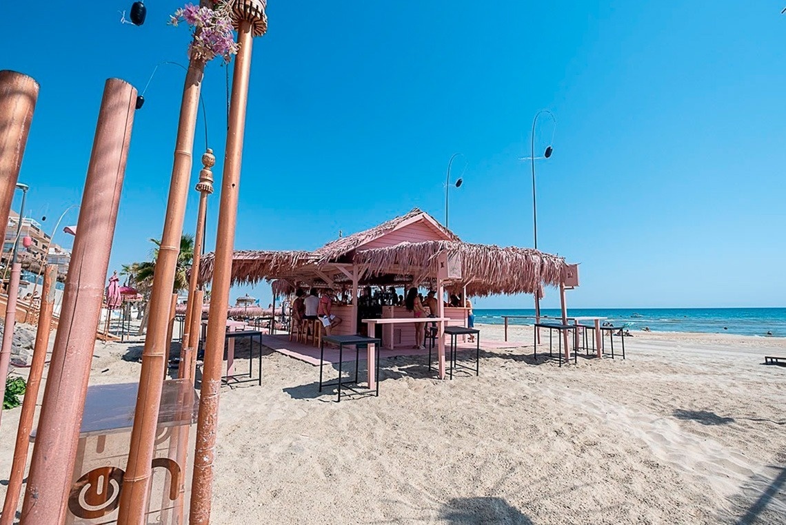 un bar rosa en la playa está rodeado de palos de bambú