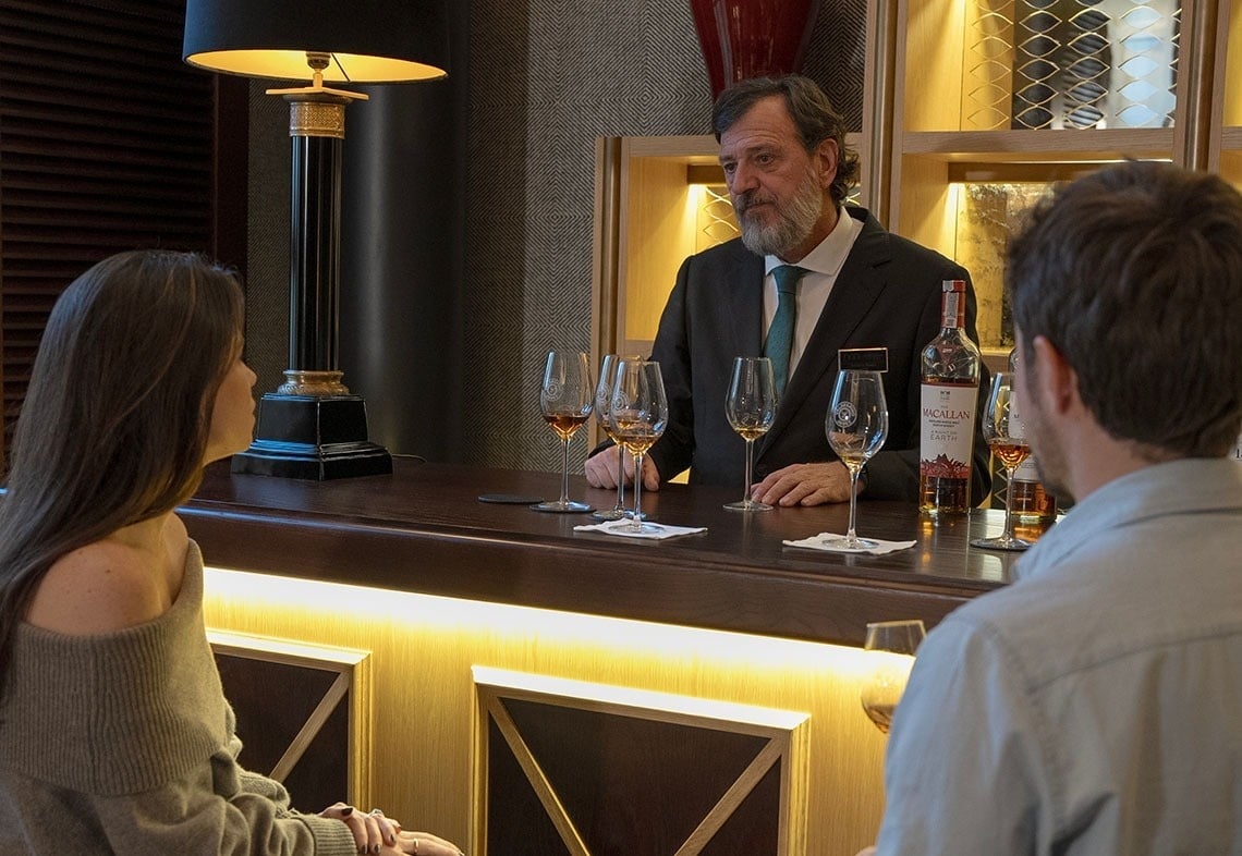 a man and a woman sit at a bar with bottles of macallan on the shelves