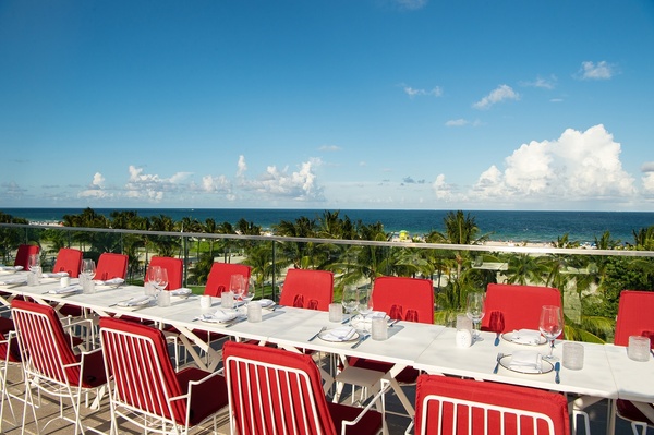Um luxuoso e moderno lobby de hotel apresenta sofás de veludo roxo, detalhes em madeira escura, plantas exuberantes e um bar elegante ao fundo.