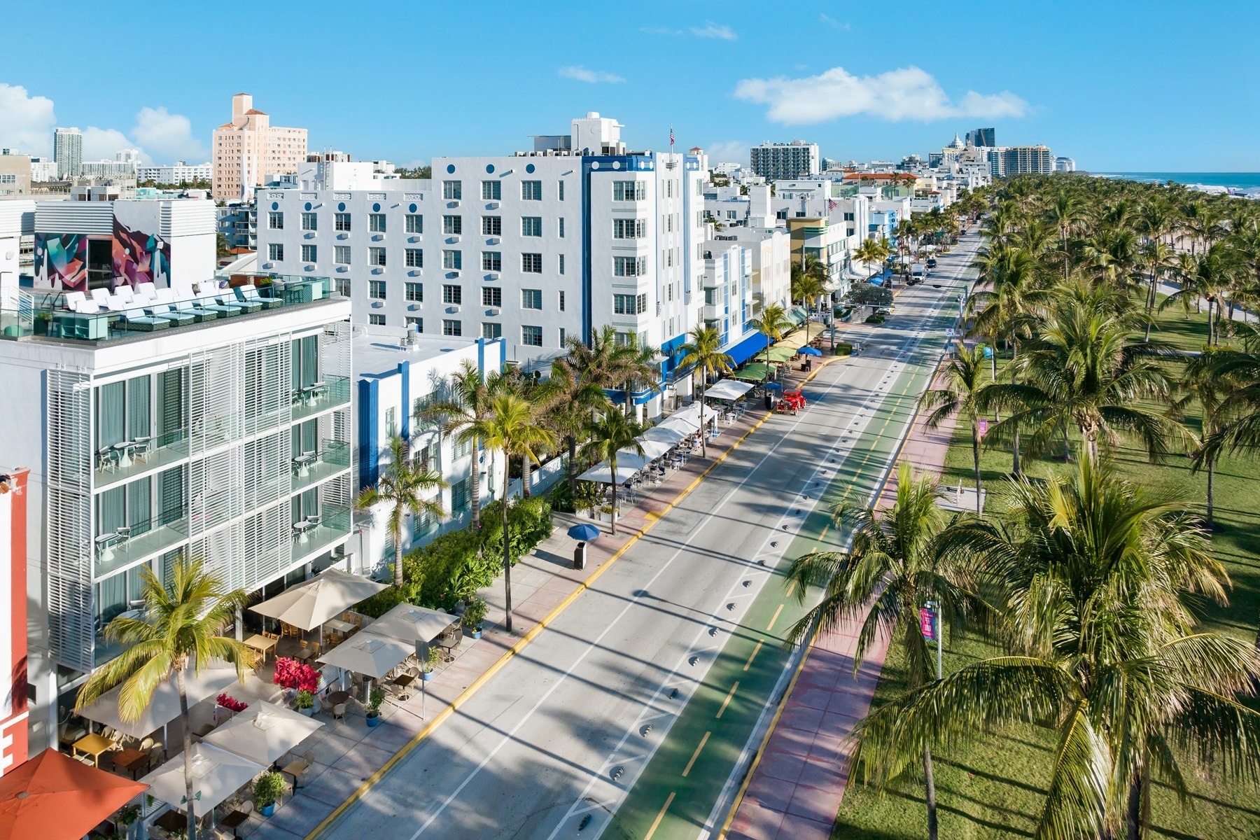 An aerial view captures the lively Art Deco hotels and outdoor cafes lining Ocean Drive in Miami Beach, with palm trees and the ocean visible in the background under a clear blue sky.