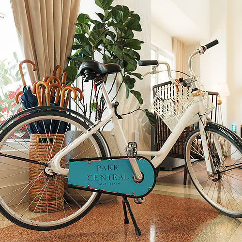 Bicicleta blanca para huéspedes con canasta y logo REEM: Park Central South Beach Hotel, junto a paraguas y planta.