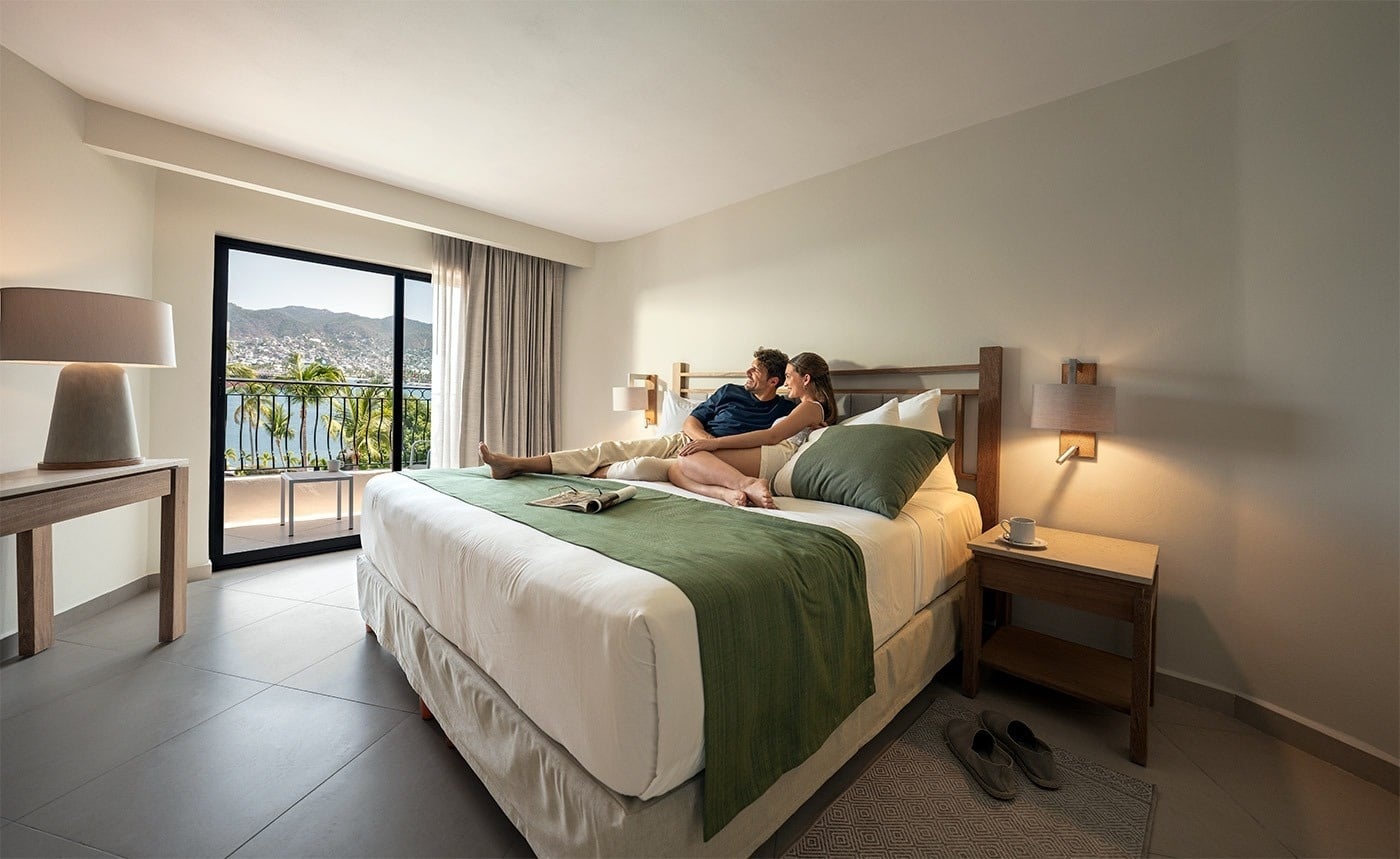 Pareja relajándose en la cama del Hotel Park Royal Beach Acapulco con vistas al Pacífico Mexicano.