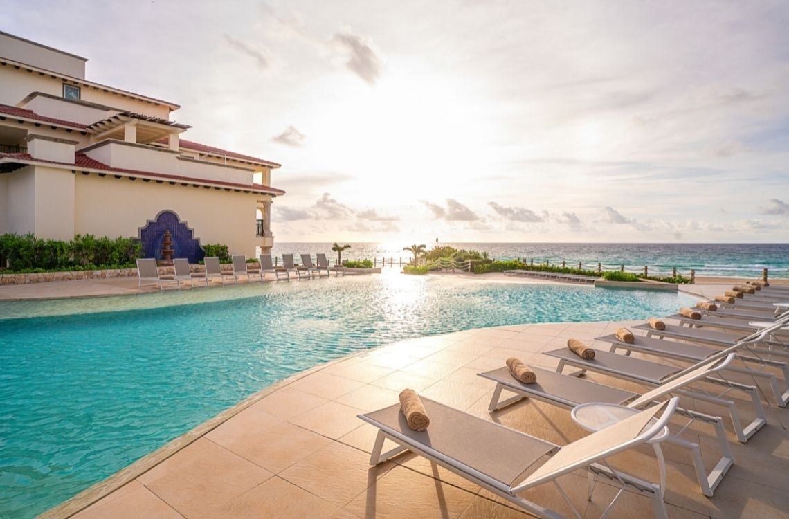 La imagen muestra una piscina infinita de un resort con tumbonas frente al océano bajo un cielo brillante.