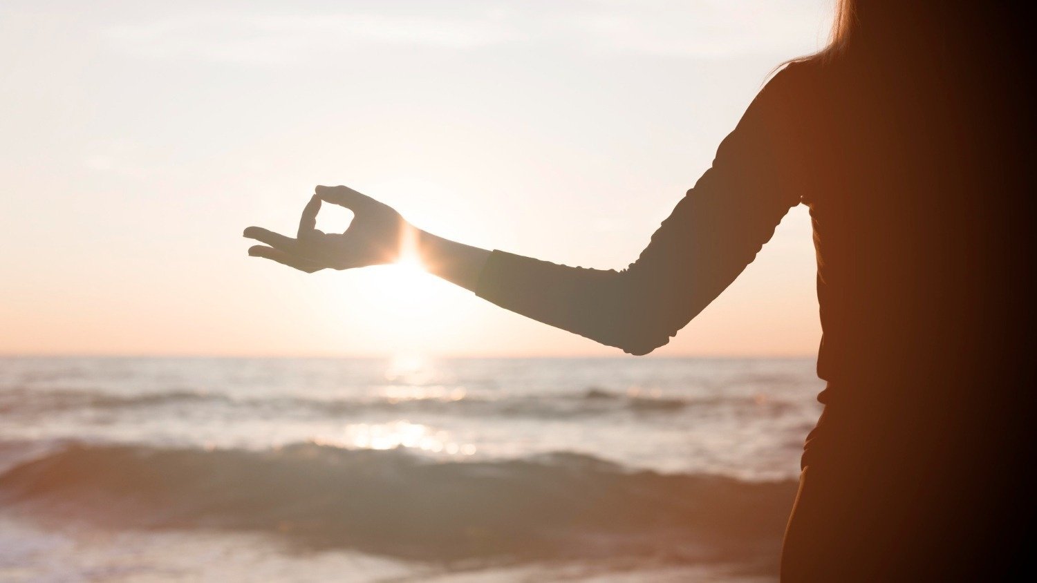 una mujer medita en la playa al atardecer mientras el sol brilla a través de sus manos