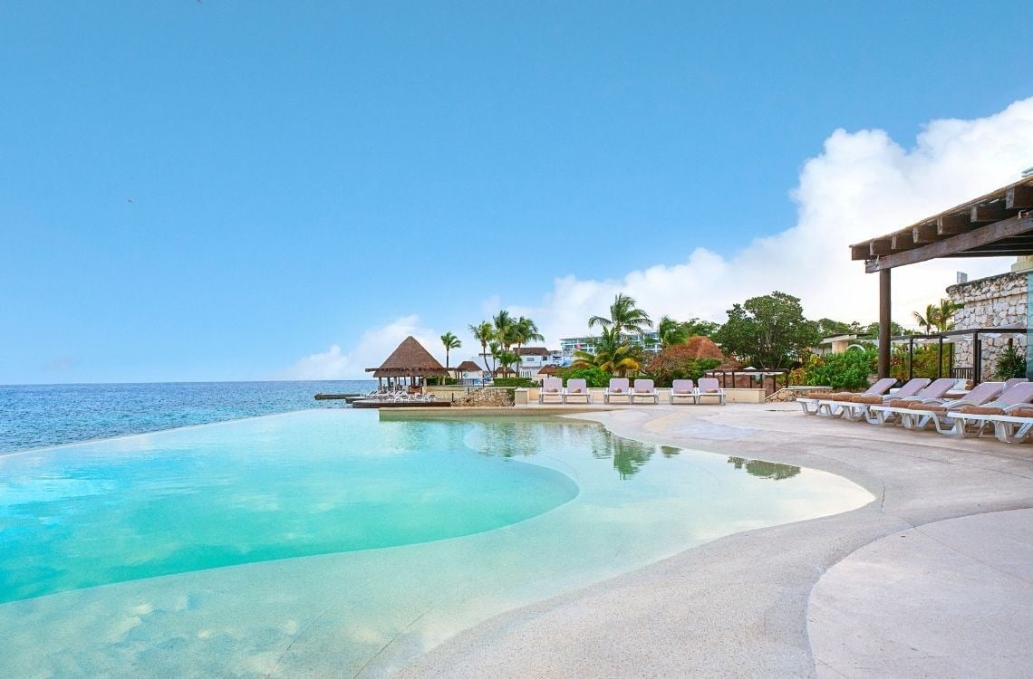 Una piscina infinita de color turquesa se fusiona con el mar, rodeada por un complejo turístico tropical con palmeras y tumbonas bajo un cielo azul claro.
