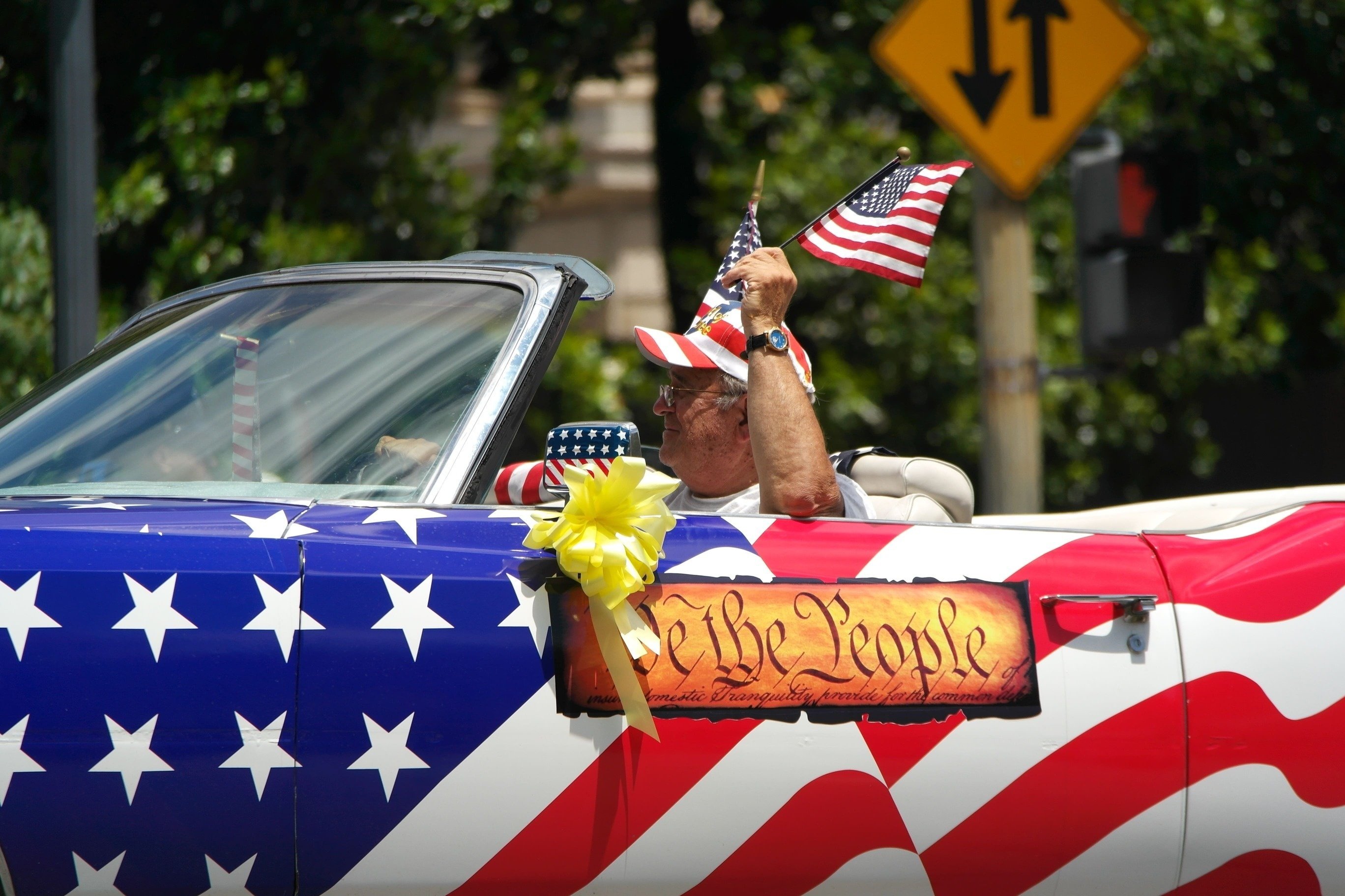 un hombre sostiene una bandera americana mientras conduce un descapotable decorado con banderas americanas
