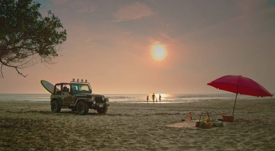 Família fazendo piquenique na praia ao pôr do sol, com jipe e prancha de surf no Park Royal Beach Ixtapa Hotel