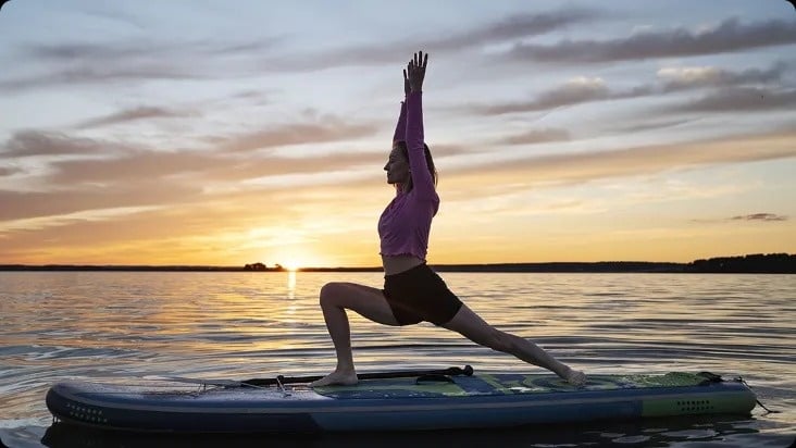 uma mulher está praticando ioga em uma prancha de surf no lago ao pôr do sol .