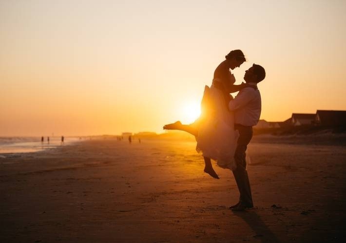 un novio sostiene a su novia en sus brazos en la playa al atardecer .