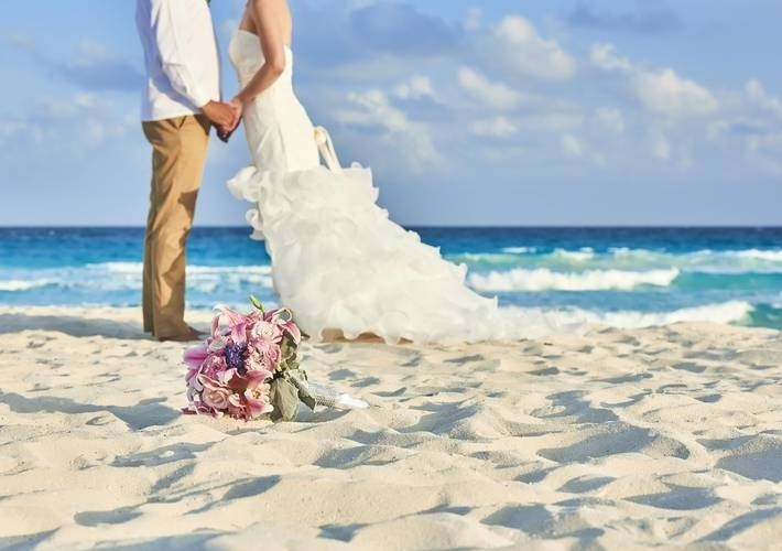 uma noiva e um noivo estão de pé na praia com um buquê de flores na areia .