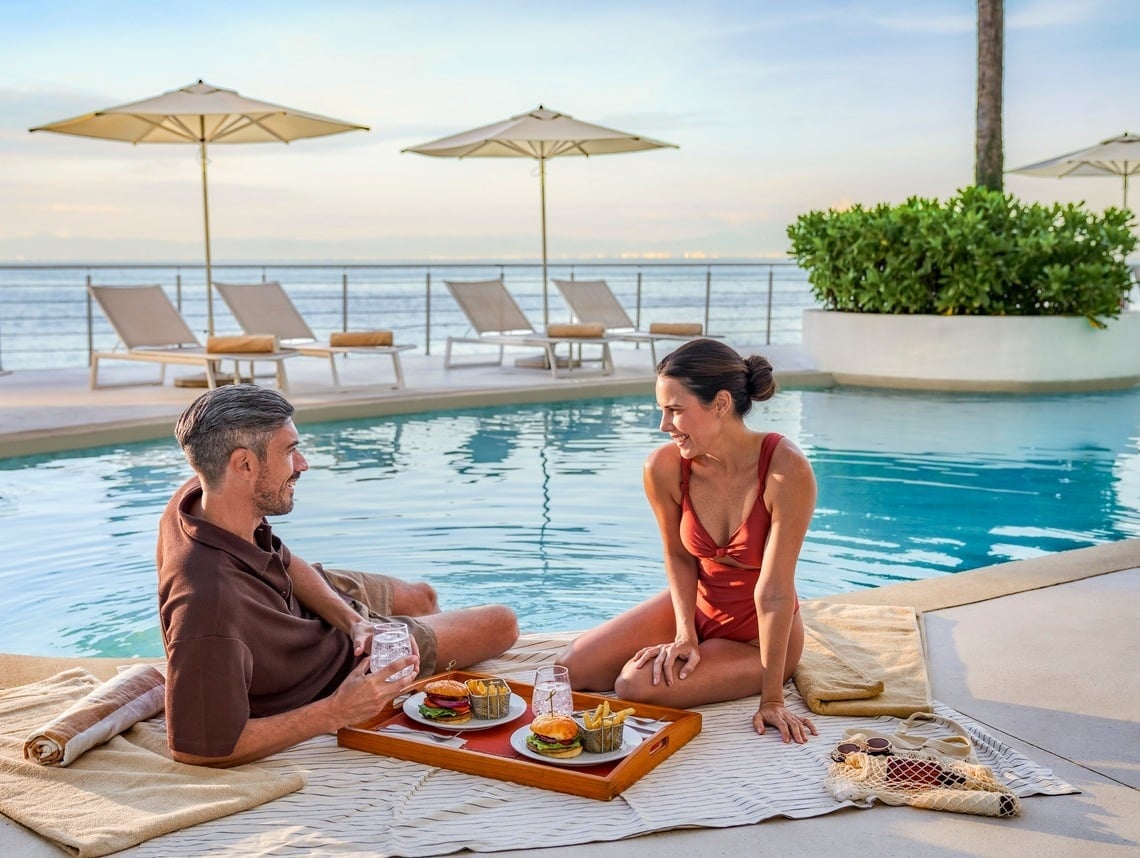 a man and woman sit on a blanket by a pool with a tray of food