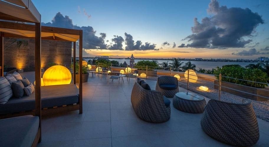 Área de relaxamento com cama balinesa, mesas, luzes e praia ao pôr do sol do Grand Park Royal Cancun Hotel
