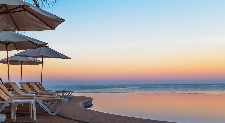 una piscina infinita rodeada de sillas y sombrillas con vistas al océano al atardecer