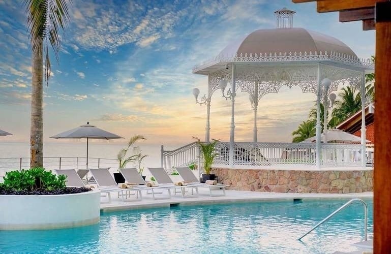 um gazebo branco ao lado de uma piscina com cadeiras e guarda-chuvas