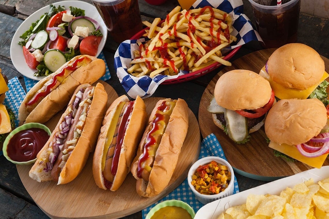 una mesa llena de comida incluyendo perros calientes hamburguesas y papas fritas