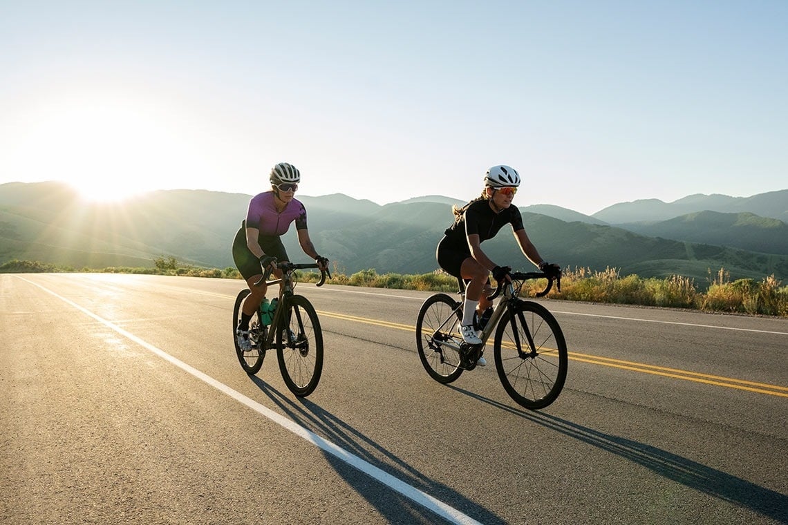 two people riding bicycles down a road with mountains in the background